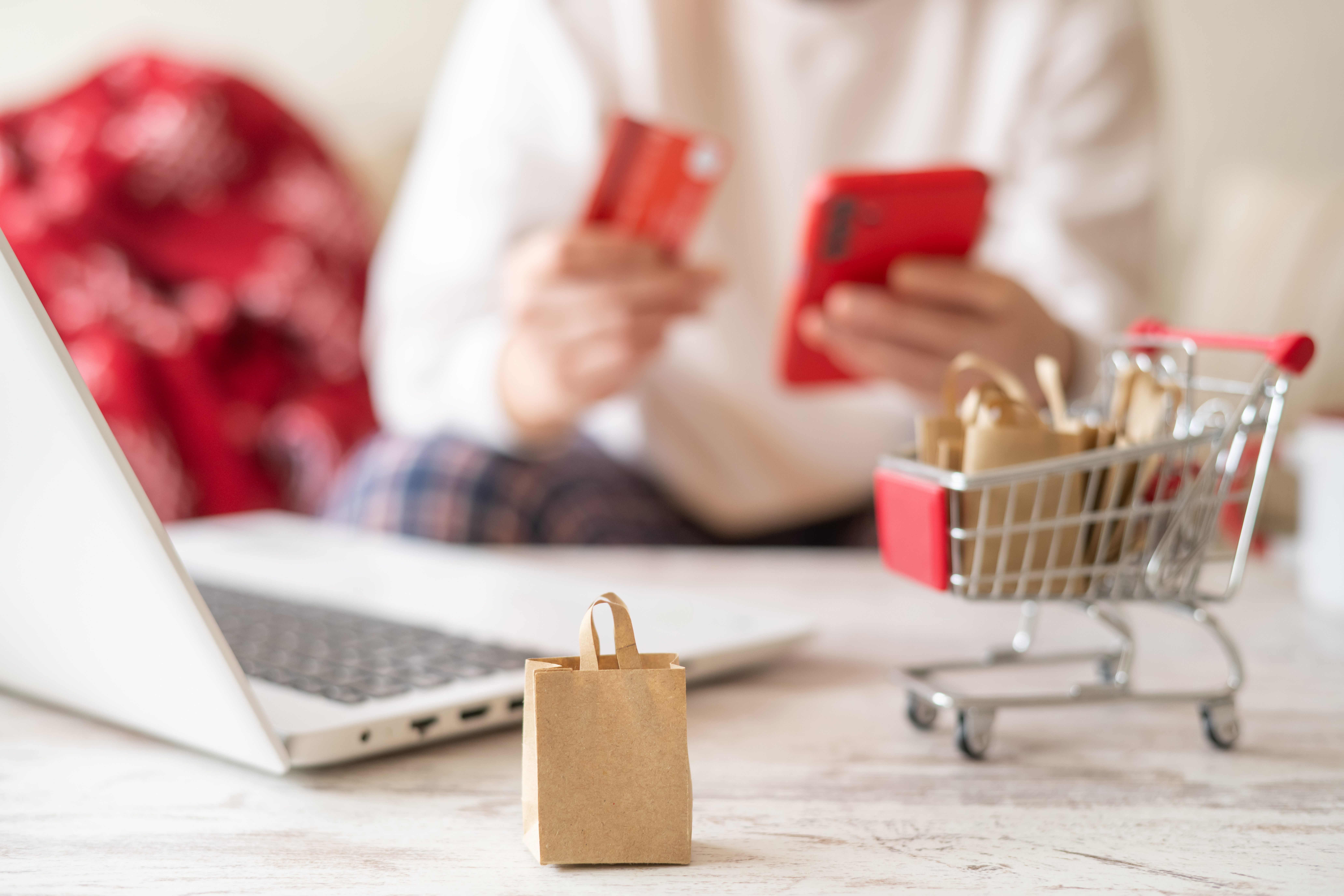Miniature shopping cart, paper bags and laptop on the foreground, while a woman is holding a credit card and using a smartphone for online shopping on a defocused background
