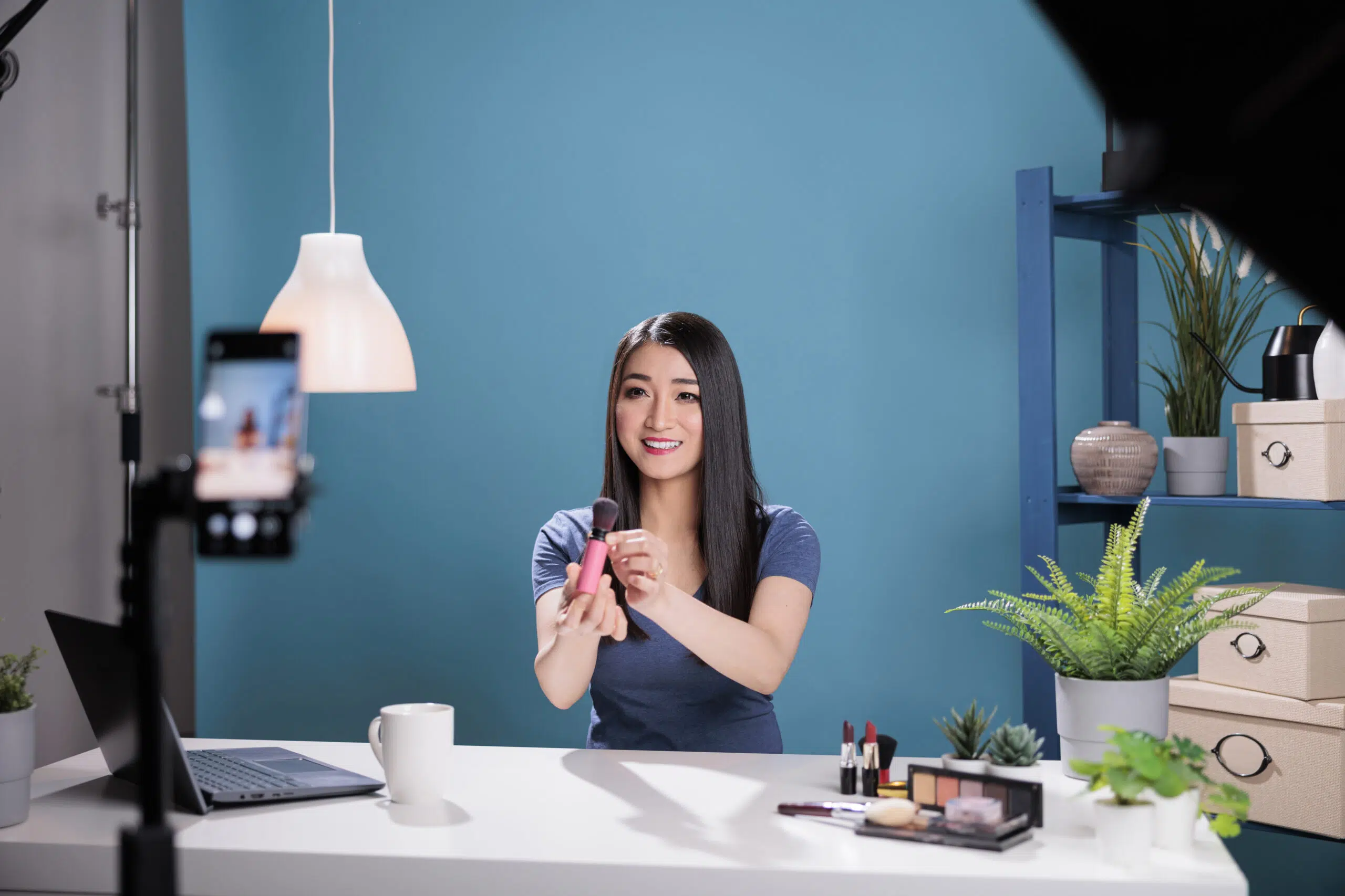 Behind the scene of asian influencer filming make up brush review using smartphone camera in studio with blue background. Vlogger creator recording cosmetics content advertising makeup product