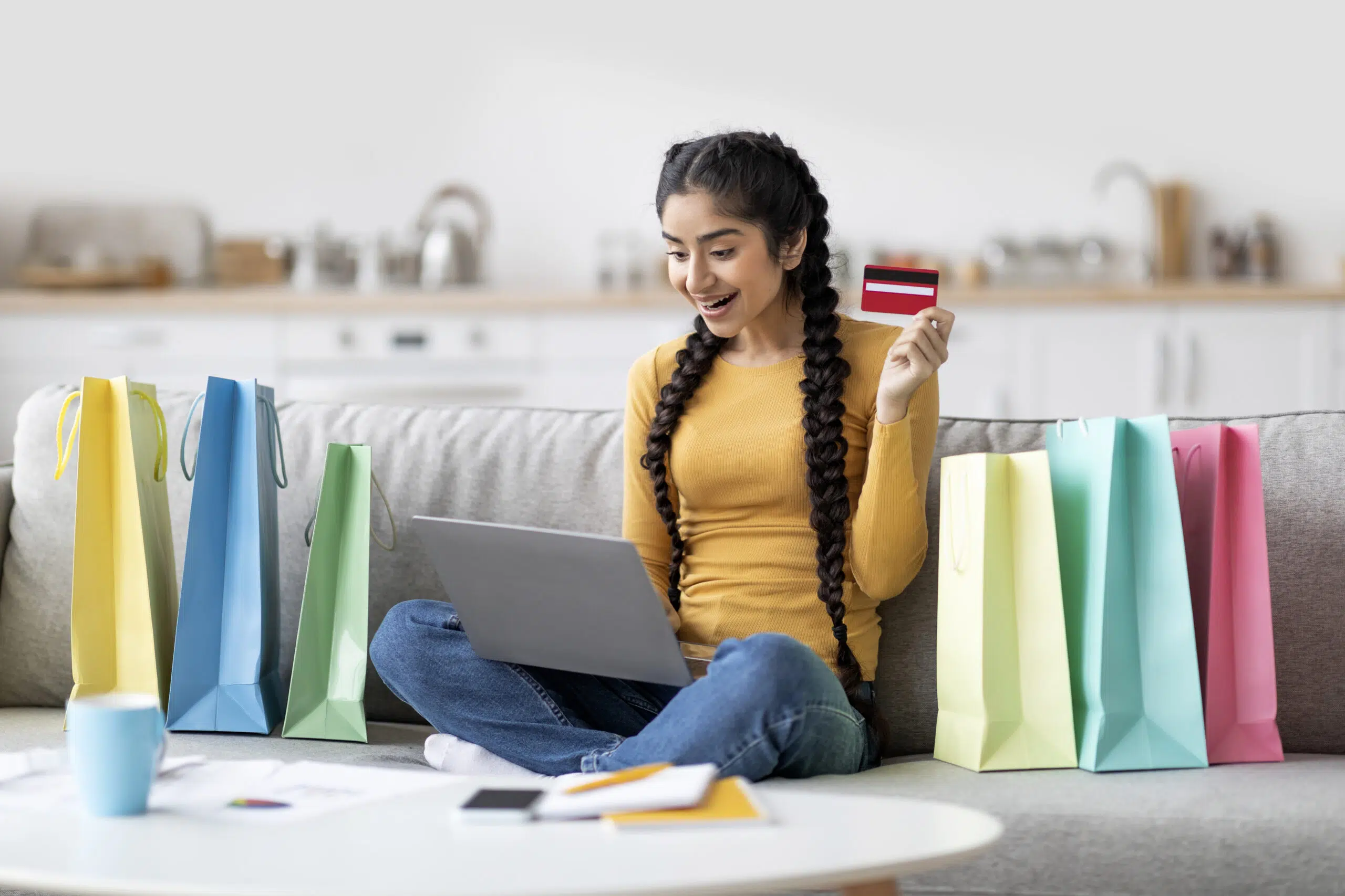Online Shopping Concept. Excited Indian Woman Using Laptop And Credit Card At Home, Happy Eastern Female Sitting On Couch With Bright Shopper Bags, Enjoying Making Pucrhases In Internet, Free Space